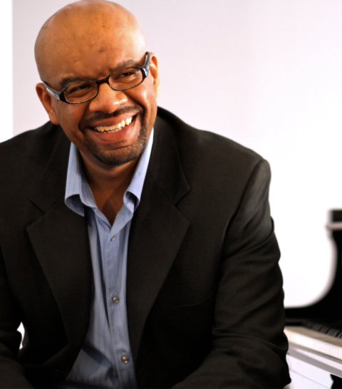 Luis Perdomo sitting in front of a piano in a black suit and blue collared shirt smiling